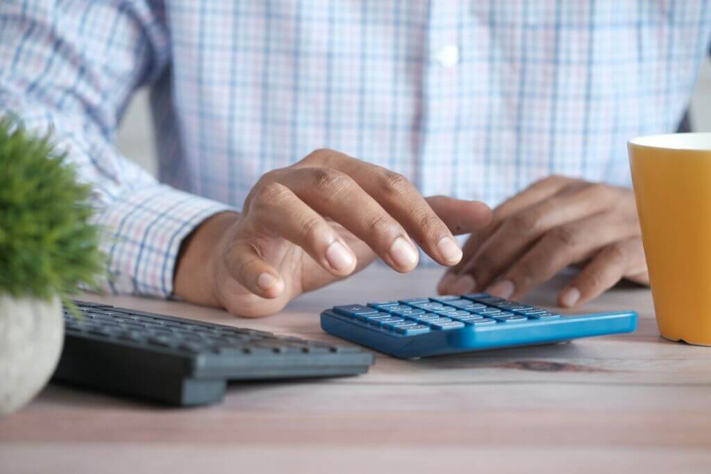 Hands using a calculator on a desk, with a keyboard and plant nearby.