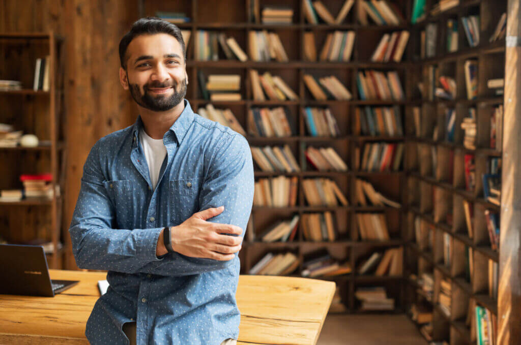 Smiling,Bearded,Indian,Businessman,Stands,Near,Desk,And,Looks,At