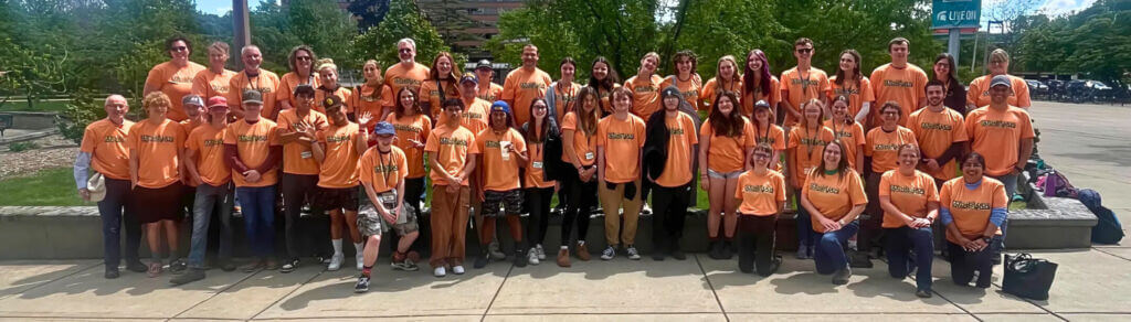 A wide panoramic group photo of high school students and coaches in orange shirts at the 2024 Michigan Envirothon state competition.
