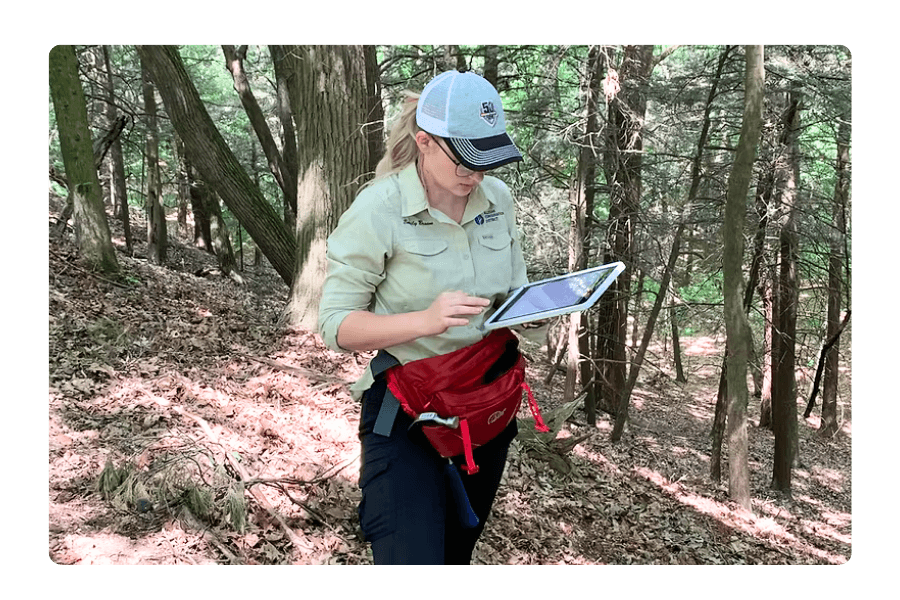 A Michigan Conservation District staff member outdoors in a wooded area, wearing a hat and branded gear, holding a clipboard and providing technical assistance.