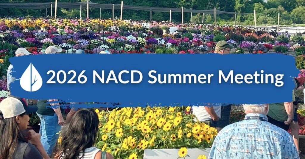 A group of people outdoors standing and walking near bright yellow and purple flowers at a nursery or garden center, overlaid with a blue paint-stroke banner containing the white Michigan Association of Conservation Districts leaf icon and the text "2026 NACD Summer Meeting".