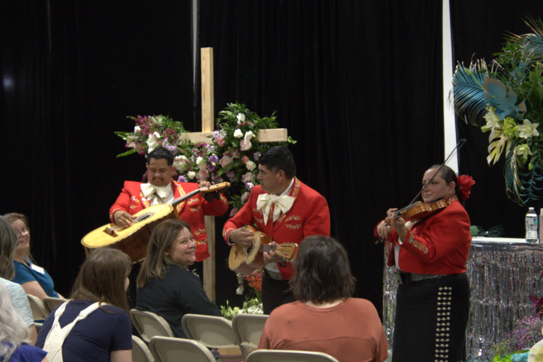 Mariachis play in front of a cross draped in florals