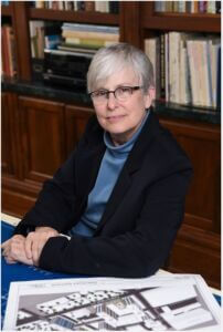 Gina Hilberry Headshot Gina is wearing a black blazer over a blue turtleneck top and is sitting at a desk with a diagram in front of her