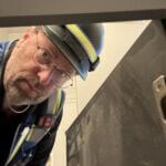 John is wearing a construction hat and a safety vest and is shown looking into a cabinet