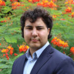 Asa Meyer Asa has dark curly hair and is wearing a blue suit. He's standing in front of a flowering bush.