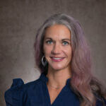 Elaine Andersen Elaine is smiling and has long hair and wearing a blue shirt.