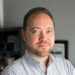 Mark Hulme Mark has short dark hair and a beard and mustache and is wearing a light blue checked shirt.