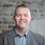 Robert Green Robert is smiling and wearing a gray suit and standing in front of a brick wall