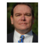 Bill is wearing a dark suit, white shirt, and blue necktie