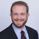 Josh is wearing a suit and tie and smiling brightly at the camera