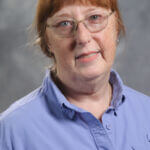 Jamie has red hair pulled back and is wearing a light purple shirt.