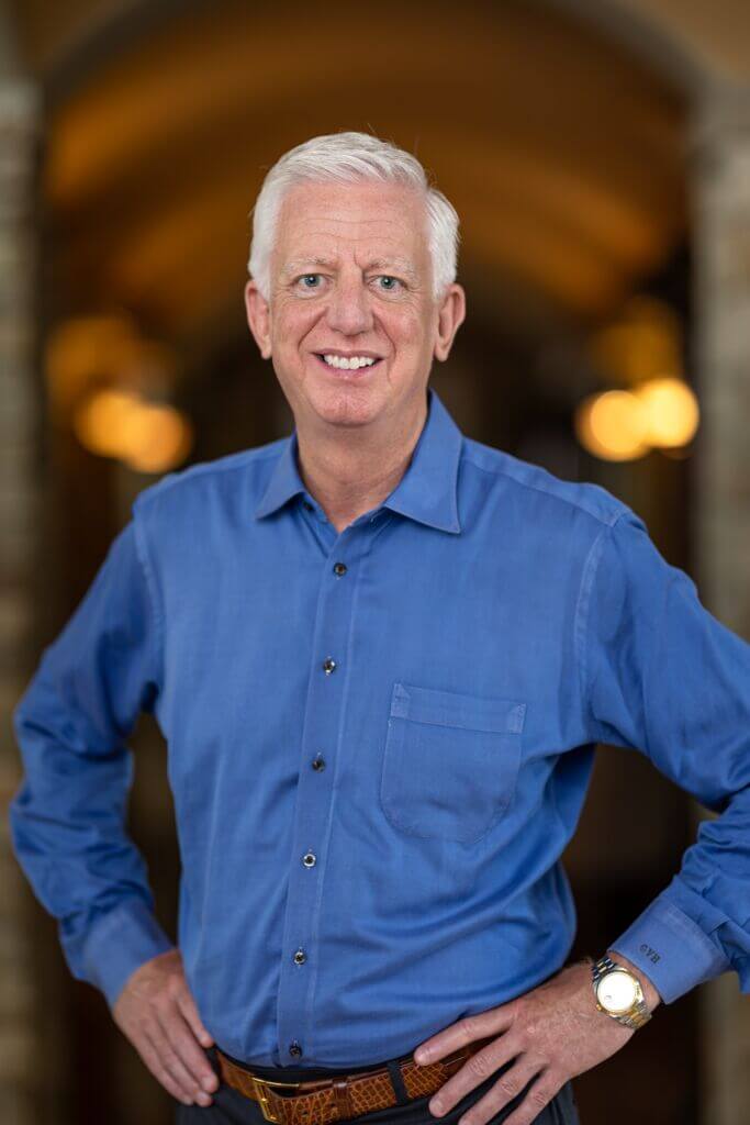 A professional headshot of a smiling man with short white hair, wearing a blue button-down shirt and a gold watch, standing with his hands on his hips against a blurred indoor background.