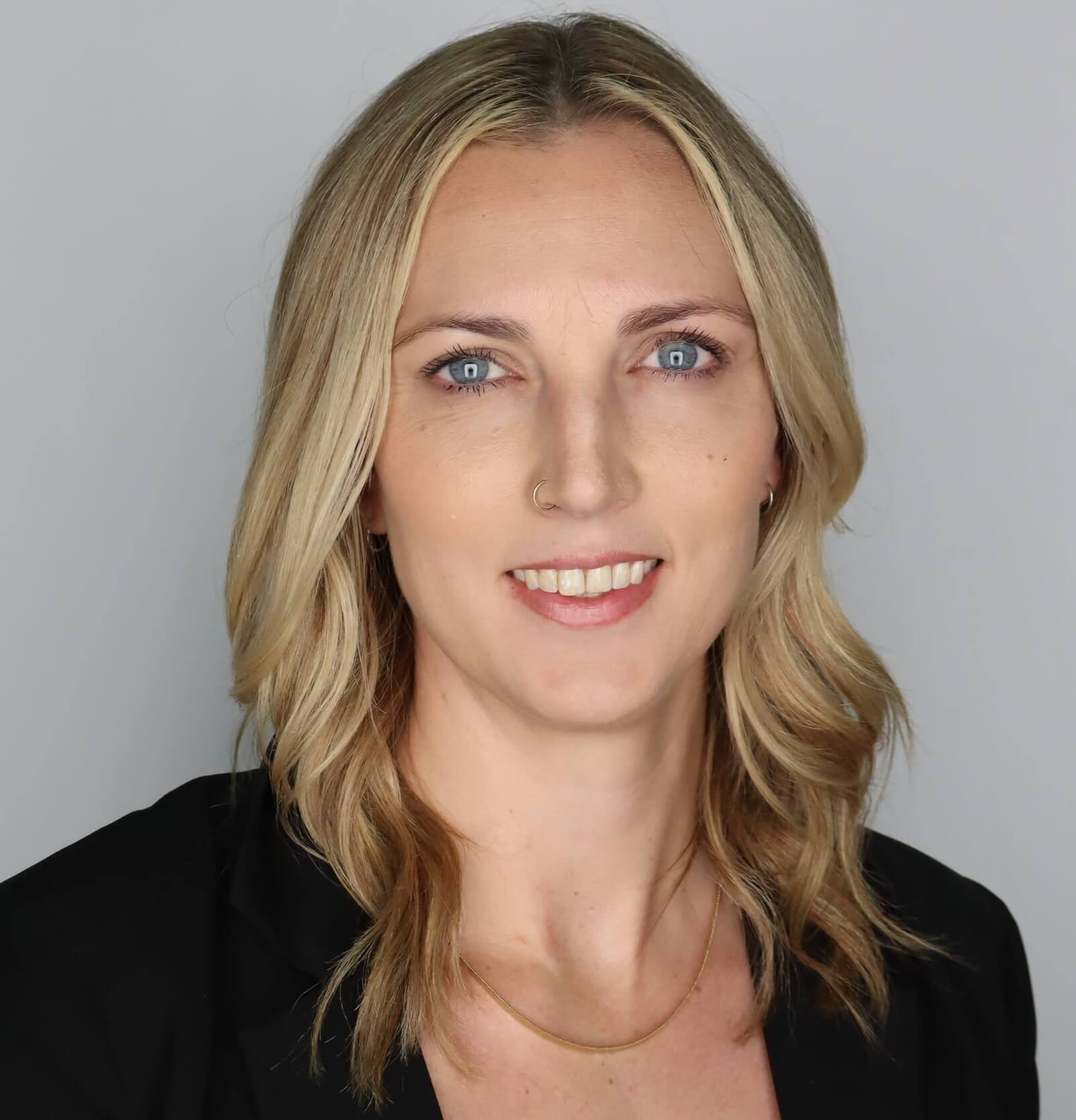 A headshot of a woman with shoulder-length blonde wavy hair and blue eyes, smiling at the camera. She is wearing a black blazer and a thin gold chain necklace. She has a small gold hoop nose ring in her right nostril and small hoop earrings. The background is a solid light gray.
