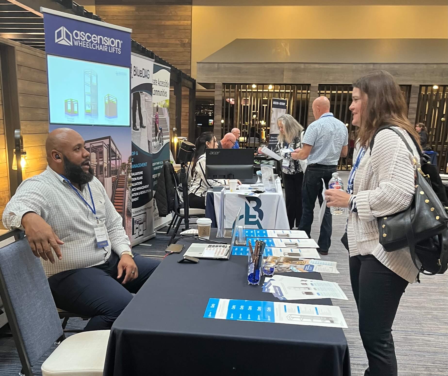 An exhibitor booth at a professional conference. On the left, a male exhibitor sits behind a draped table conversing with a female attendee standing on the right. The table is neatly arranged with a laptop, marketing brochures, and promotional pens. Behind the exhibitor stands a tall banner for "Ascension Wheelchair Lifts" featuring a digital screen with product diagrams. In the background, other attendees interact at adjacent booths, including one for "BlueDAG."