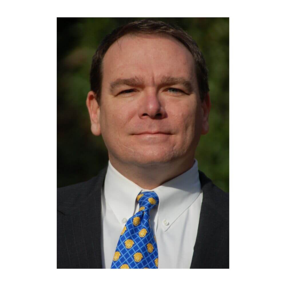 Headshot of Bill Hecker, a man with short brown hair, wearing a dark suit jacket, white shirt, and a blue tie with a gold pattern.