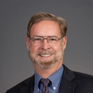 Professional headshot of Bill Zellmer, a smiling man with a beard and glasses, wearing a suit jacket, a blue patterned button-down shirt, and a tie against a solid gray background.