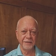Headshot of a grinning man with a white goatee and blue shirt standing in front of a brown wall.