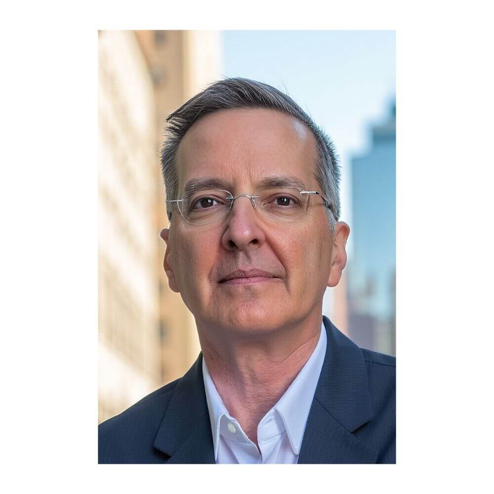 Headshot of Jim Safranek, a man with short, graying hair and rimless glasses, wearing a dark suit jacket and an open-collar white shirt against a blurred city background.