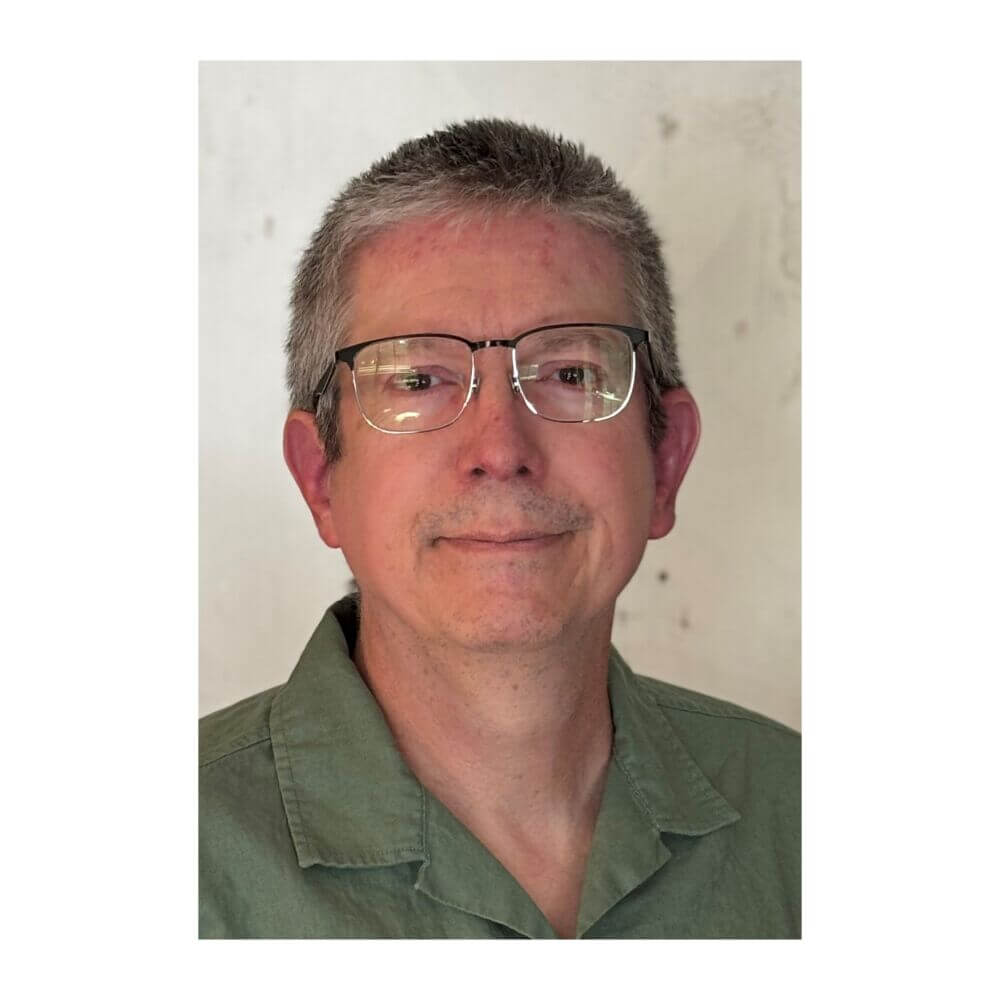 Headshot of John Gonzalez, a man with short graying hair and glasses, wearing a green collared shirt against a plain, light background.