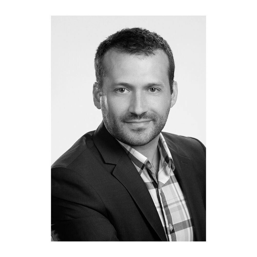 Black and white headshot of Jonathan White, a man with short hair and light stubble, wearing a suit jacket and a plaid button-down shirt.