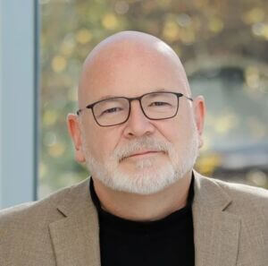 Headshot of Joseph Cooley, with a short, trimmed white beard and mustache, wearing black-rimmed glasses, a black turtleneck, and a light tan blazer against a softly blurred outdoor background.