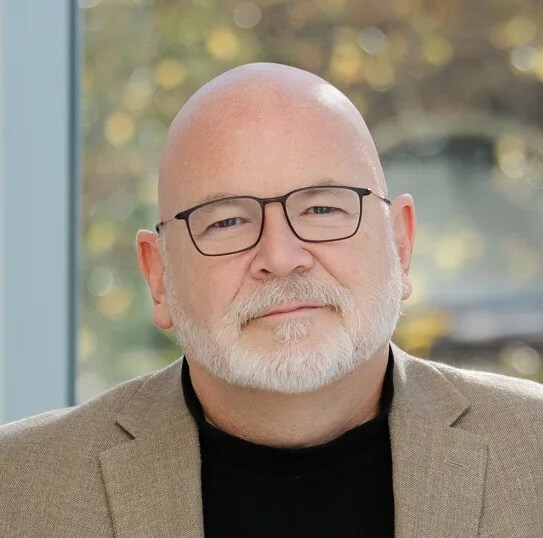 Headshot of Joseph Cooley, with a short, trimmed white beard and mustache, wearing black-rimmed glasses, a black turtleneck, and a light tan blazer against a softly blurred outdoor background.