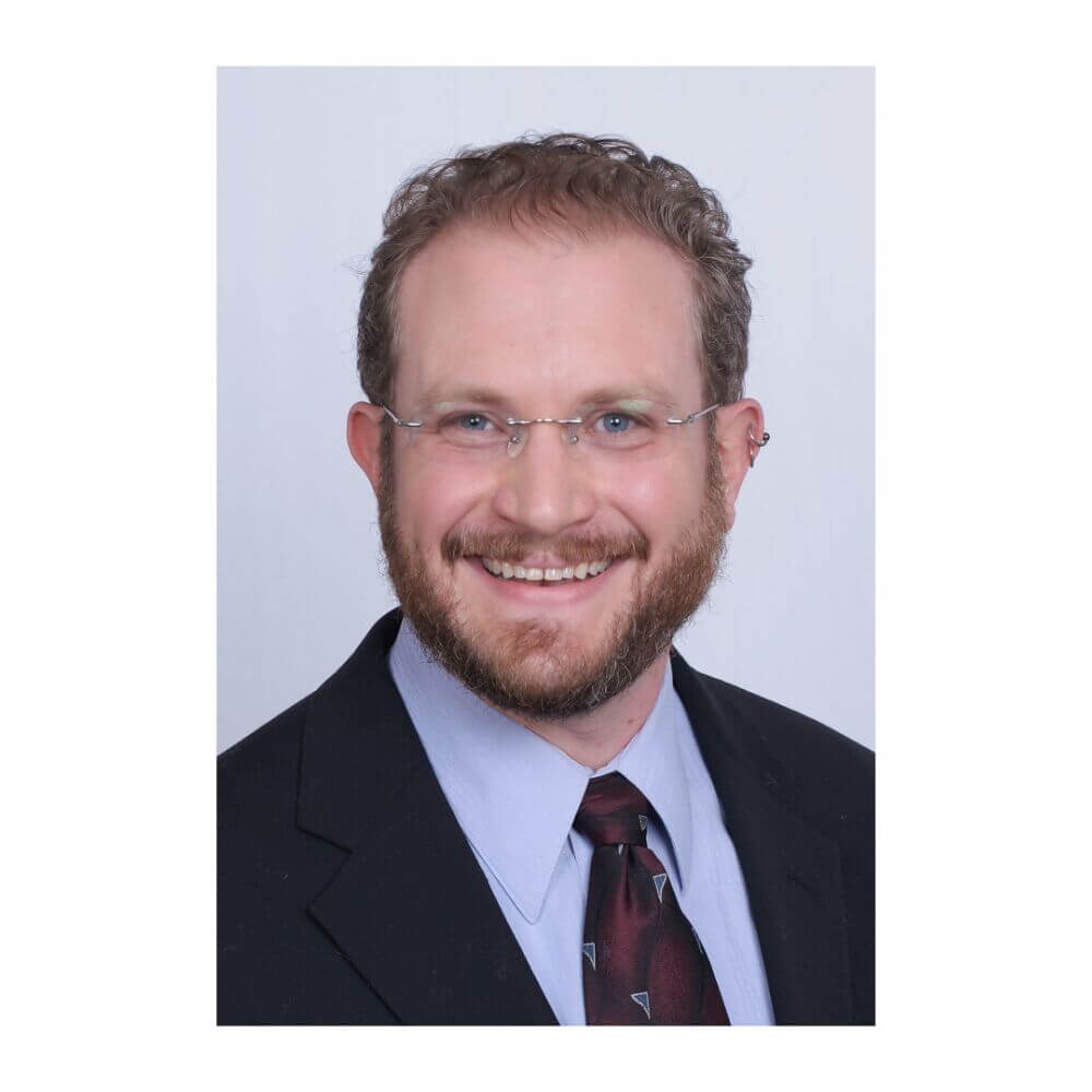 Headshot of Josh Schorr, a smiling man with curly brown hair, a beard, and glasses, wearing a black suit jacket, blue shirt, and dark tie against a light gray background.