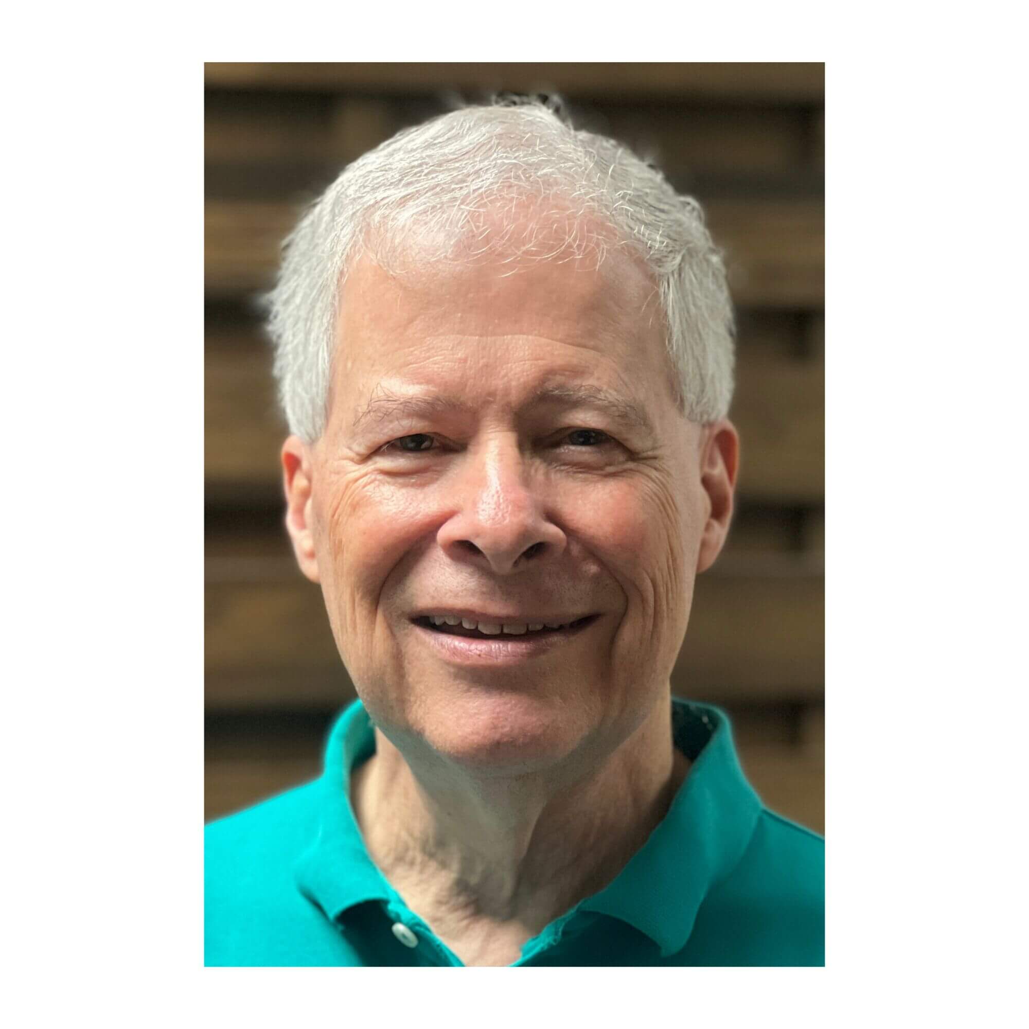 Headshot of James Terry, a man with short white hair, smiling and wearing a bright teal polo shirt against a blurred, dark wooden background