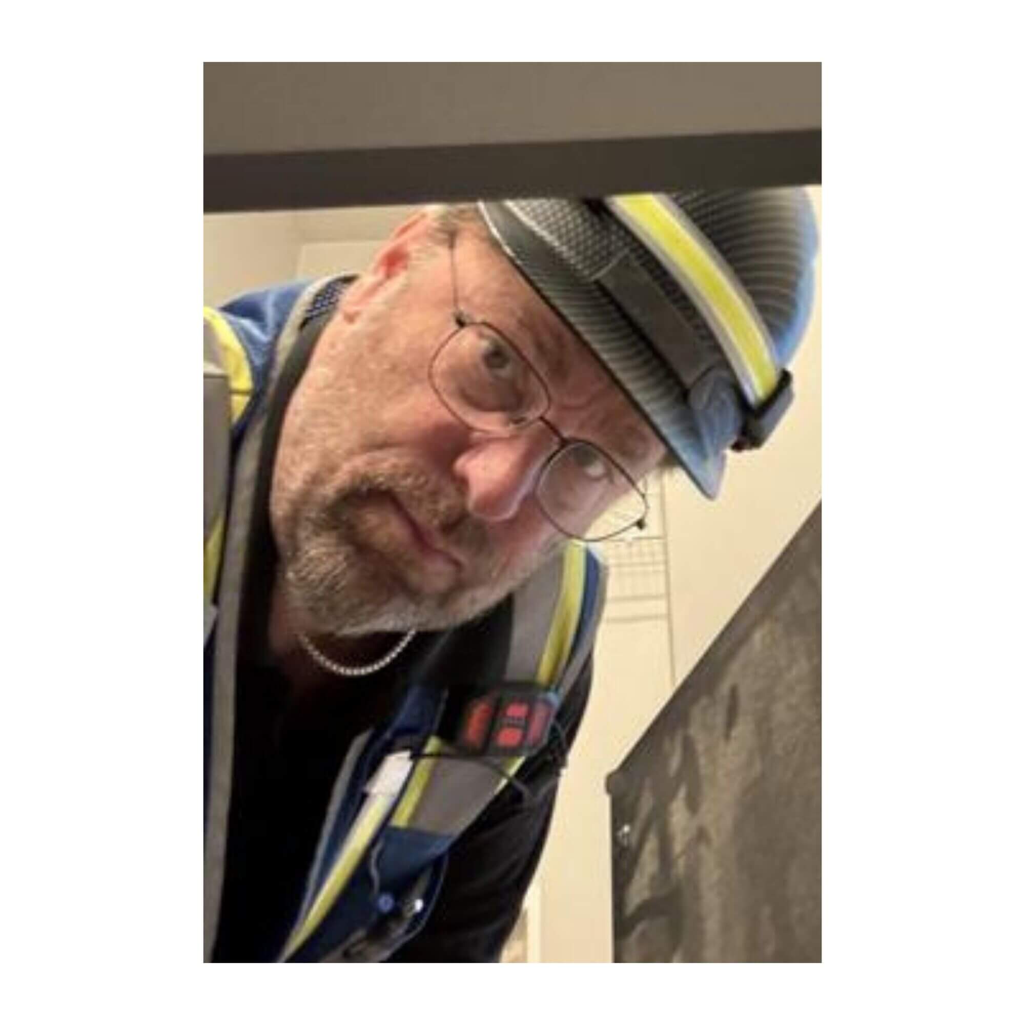 A close-up, angled photo of John Torkelson, a man with a beard, glasses, a dark hard hat with reflective striping, and a high-visibility safety vest, looking down toward the camera.