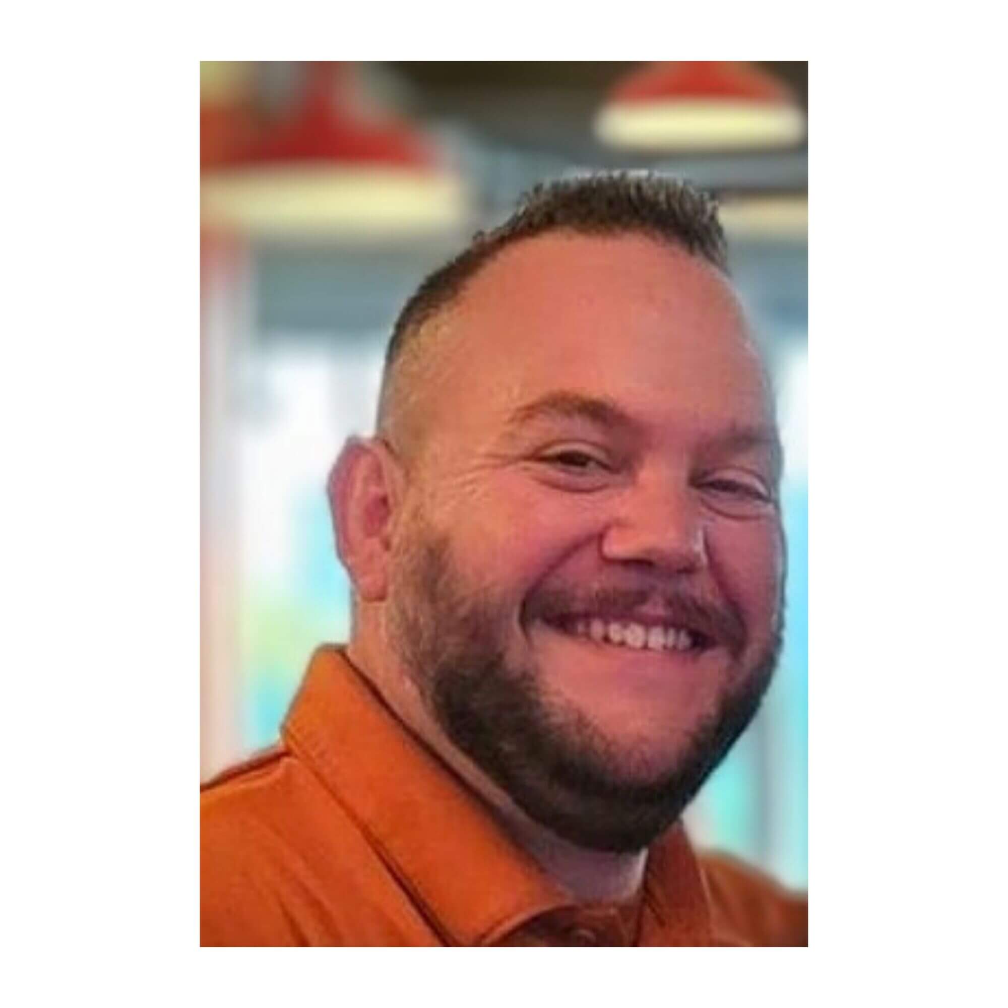 Close-up headshot of Jonathan Sparks, a man with short brown hair, a full beard, and a wide smile, wearing an orange collared shirt.