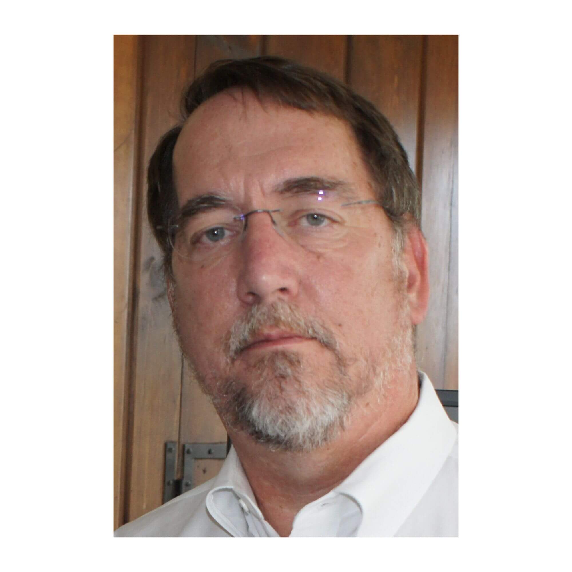 Mark Mazz (1) Headshot of Mark Mazz, a man with short brown hair, a grey-and-brown trimmed beard and mustache, and rimless eyeglasses, wearing a white collared shirt against a wooden panel background.