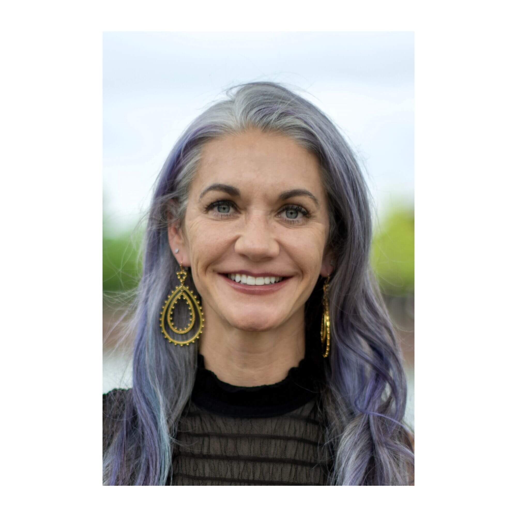 Headshot of Elaine Andersen smiling outdoors, with long silver-and-lavender hair, gold earrings, and a black high-neck top.