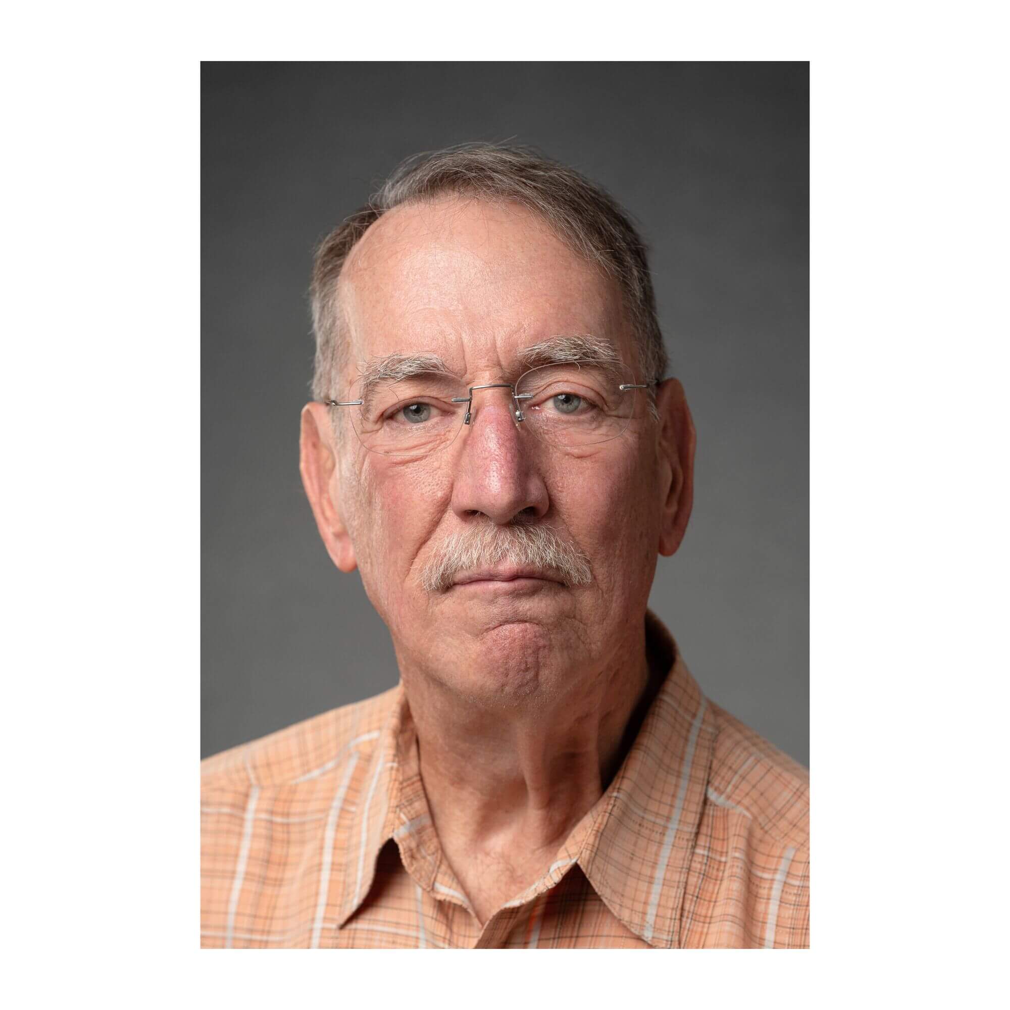 Mark Mazz Headshot of Mark Mazz, a man with short gray hair, a gray mustache, and rimless eyeglasses, wearing an orange and white plaid collared shirt against a neutral gray background.