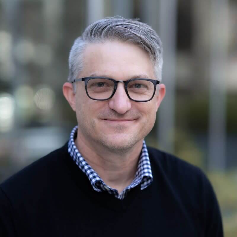 Headshot of Steven Dombrowski, a smiling man with short grey hair and black glasses, wearing a patterned shirt under a black sweater.