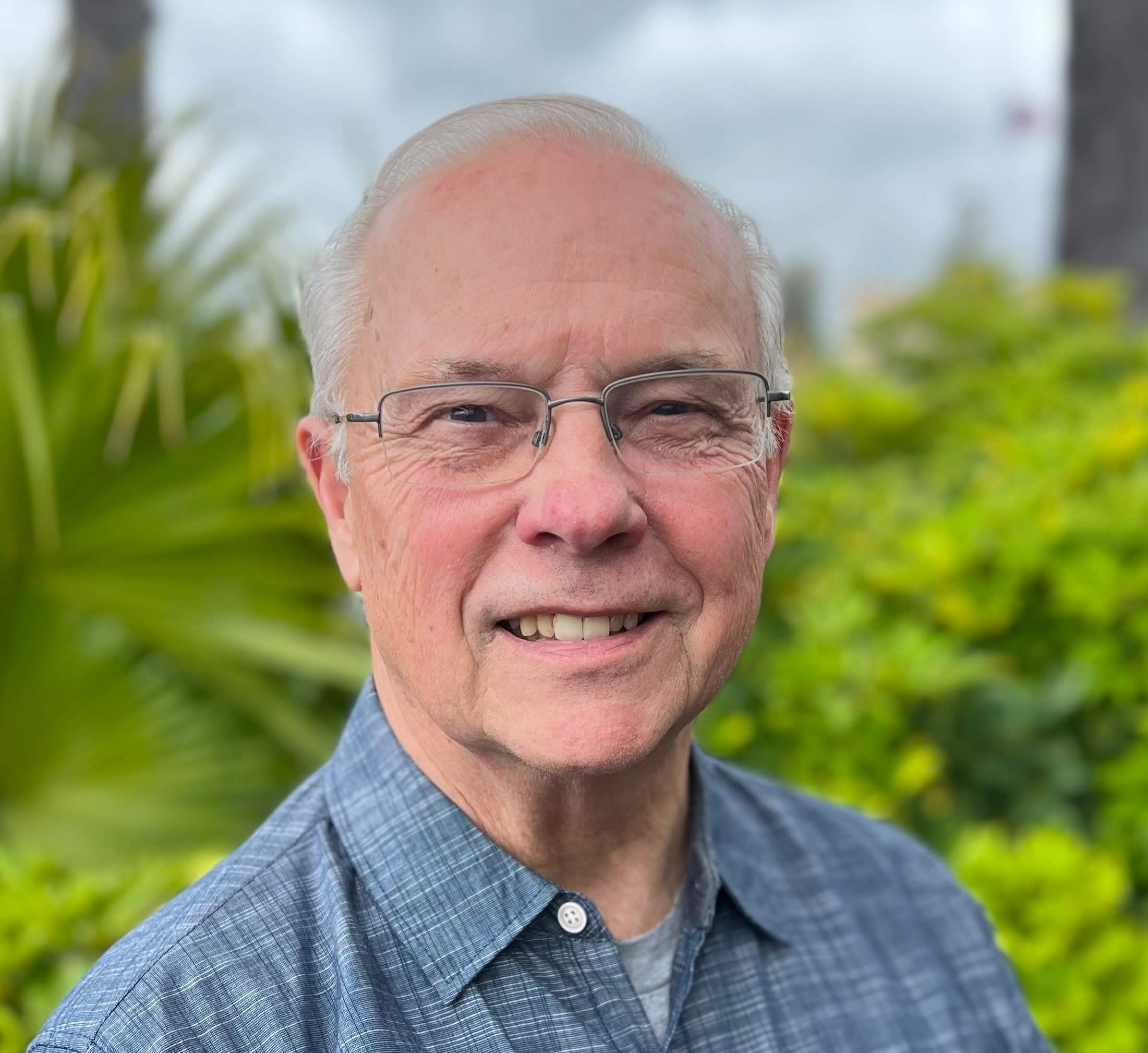Headshot of Greg Izor smiling outdoors, wearing glasses and a blue short-sleeved button-down shirt in front of green foliage.