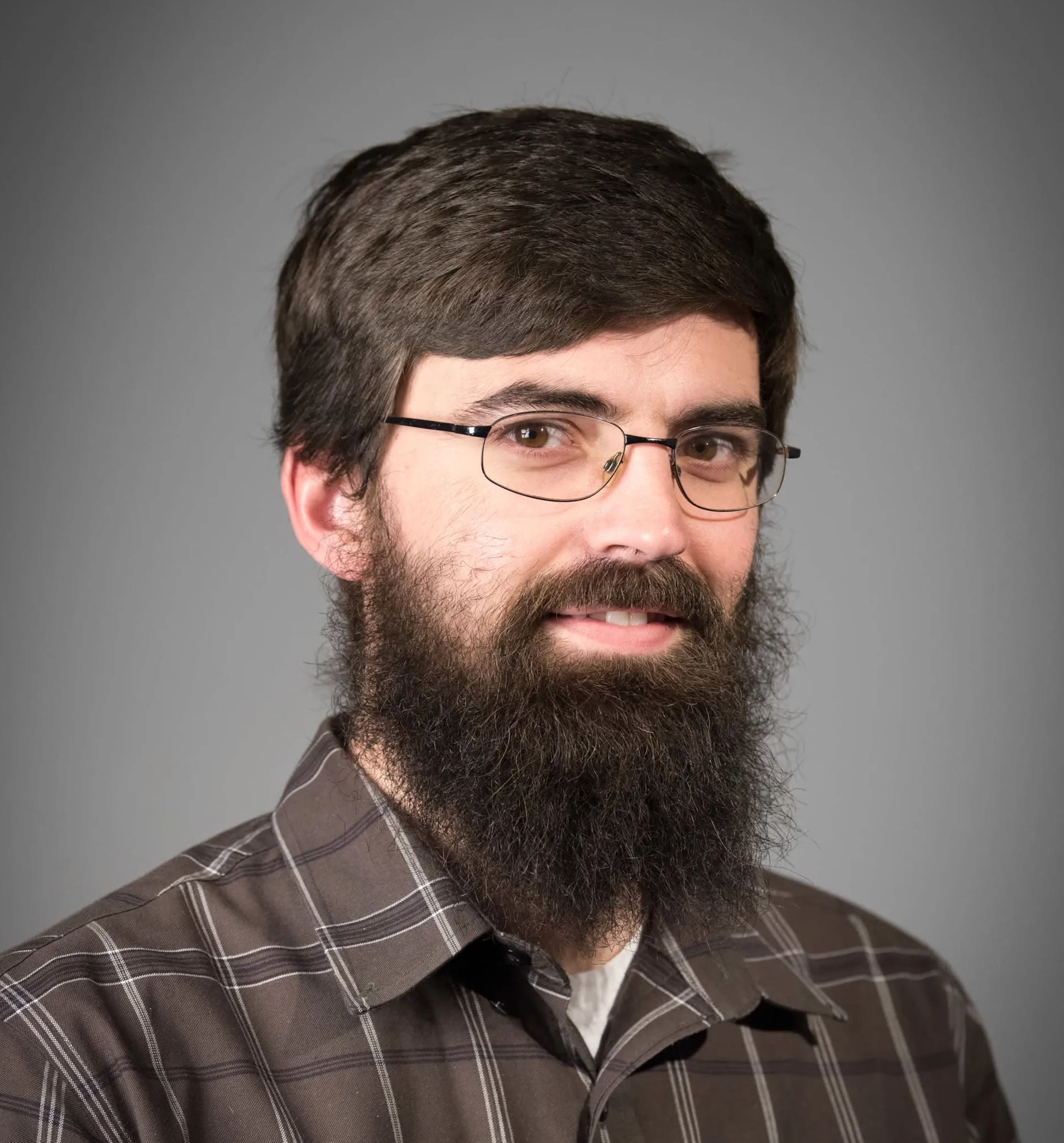 Headshot of Joshua Johnson, a man with short brown hair, a full dark beard, and wire-rimmed glasses, smiling in a brown plaid shirt against a gray background.
