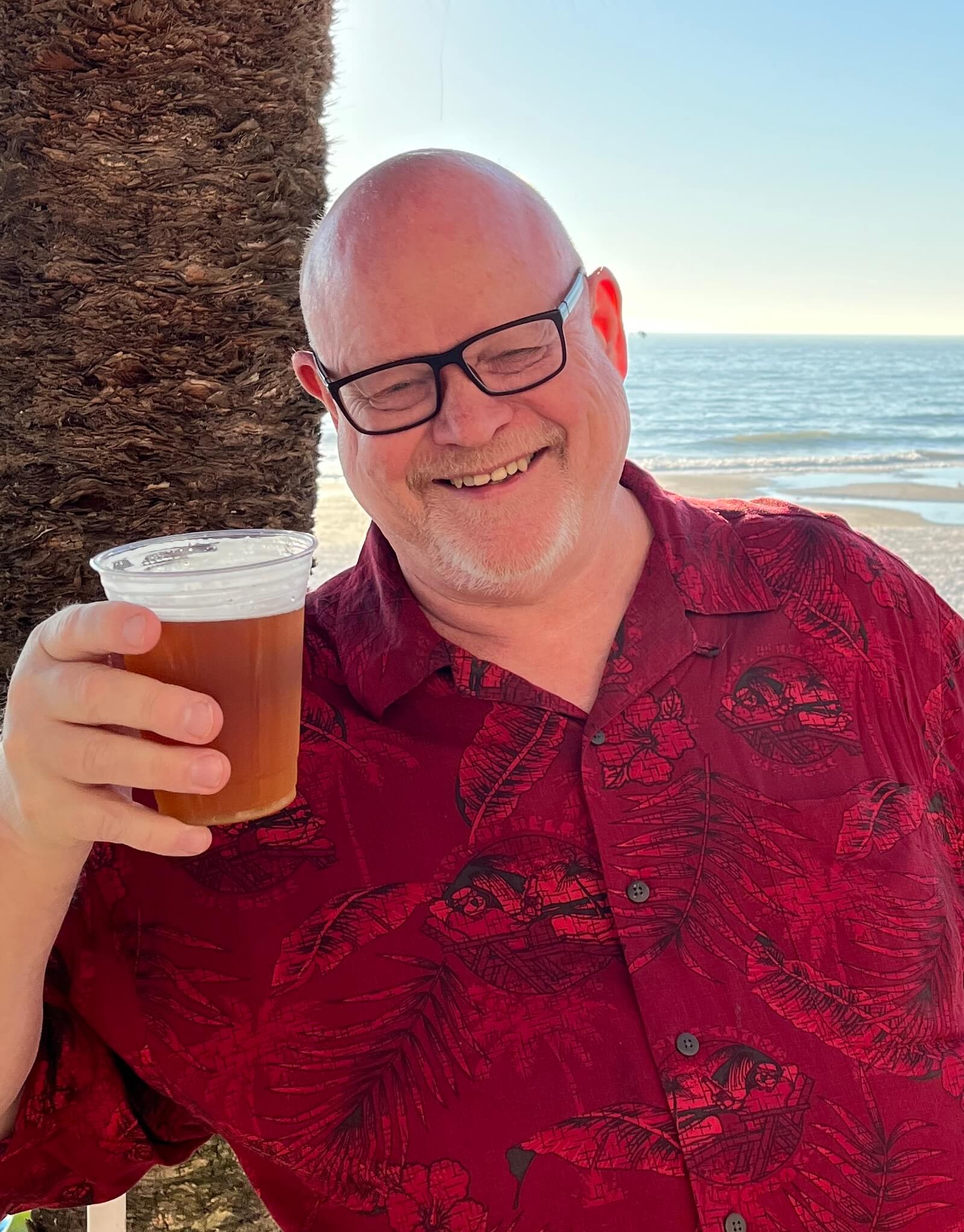 A portrait of Mark Lagerkvist holding up a beer by the beach and smiling