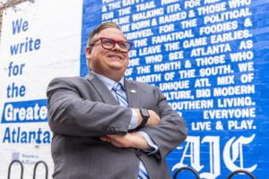 Portrait of Charles Minshew with arms crossed standing in front of a blue wall with the Atlanta Journal Constitution logo