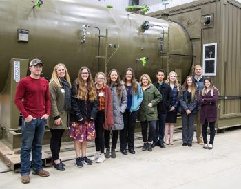 group of students and an adult posing together for a photo inside an industrial facility, with large cylindrical equipment visible behind them.