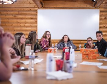 A group of young people seated around a table in a wood-paneled room, engaged in a discussion or workshop, with papers and bottles on the table and a whiteboard visible in the background.