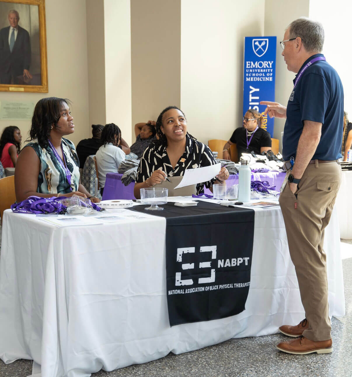 Two women seated at a National Association of Black Physical Therapists (NABPT) table speak with a man standing at a professional event.