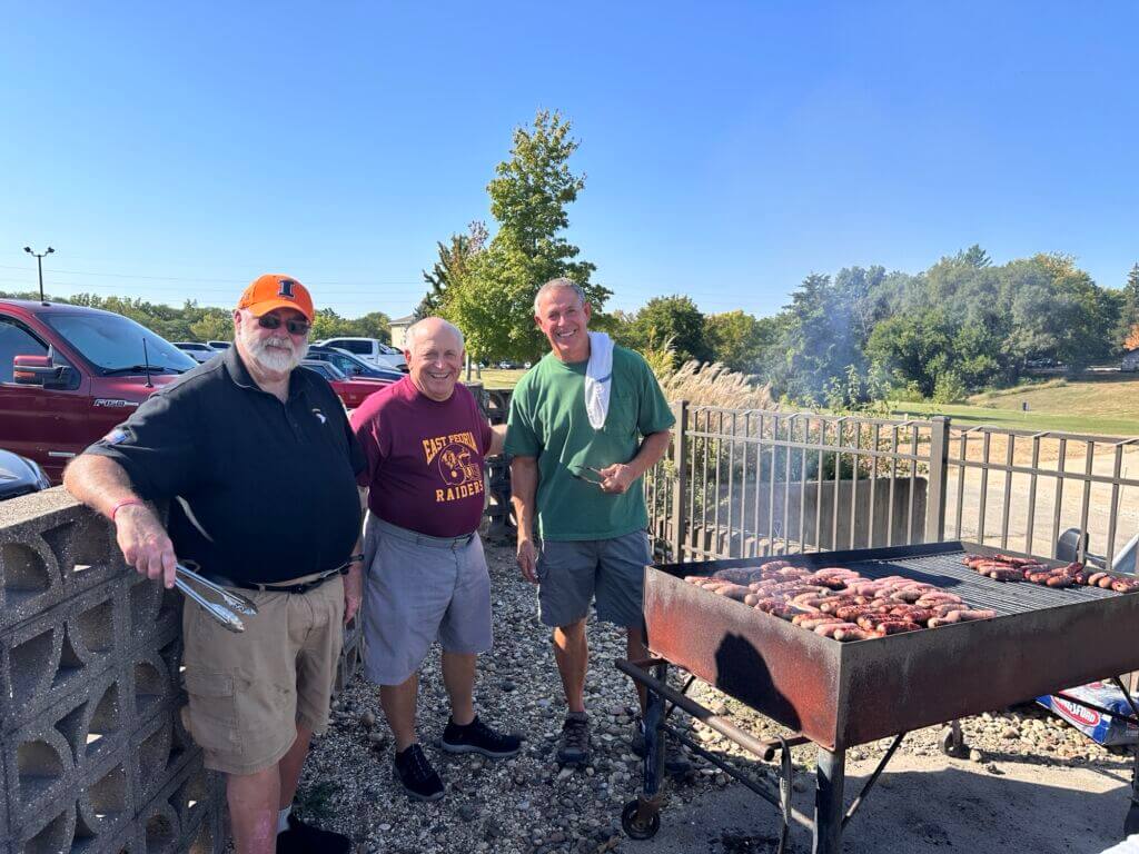 Three men stand near a smoking grill at the Golf Classic cookout