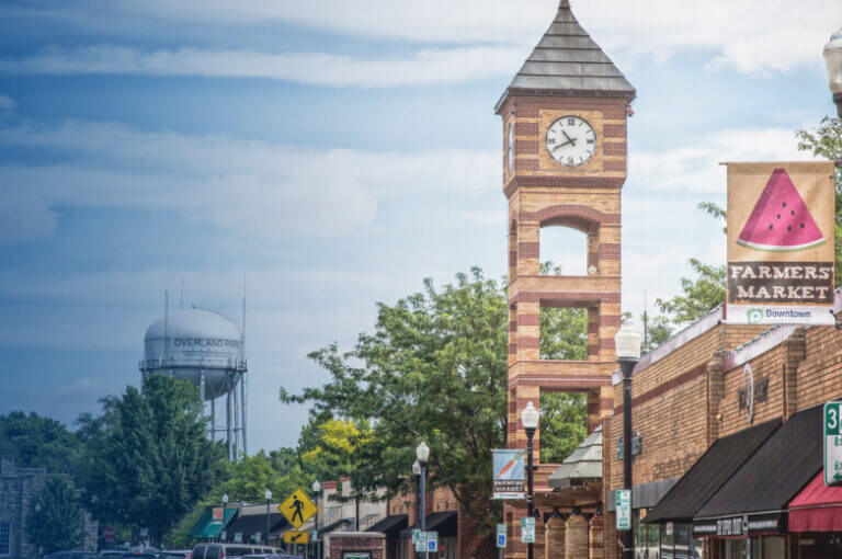 Historic brick clock tower in a downtown square surrounded by trees under a blue sky