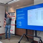 man pointing out into group of people watching his presentation with screen to the side