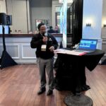 Presenter speaking into a microphone at table next to a laptop, with an event banner in the background