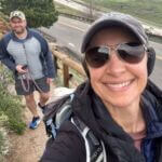 woman taking selfie while out hiking man with a dog smiling in background