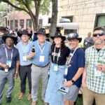group of people wearing cowboy hats standing outside with many other people standing in background networking