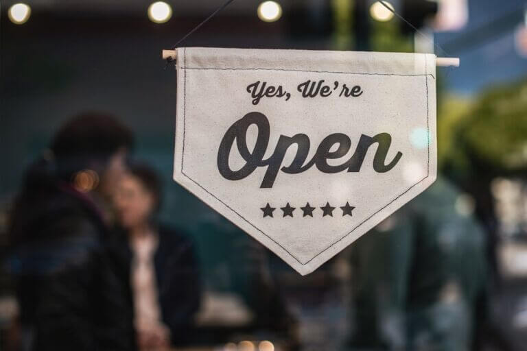 A white cloth pentagon shaped sign hanging on a glass door that reads Yes, We're Open in black text and has five stars underneath.