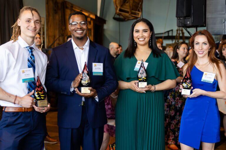 A close up showing the four winners of the Maryland Chamber of Commerce Rising Star Award, honoring young professionals, pose and hold their glass awards while a crown of onlookers stands behind them.