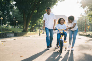 Two adults guide a child who is learning to ride a bike with training wheels on a paved road lined by trees.